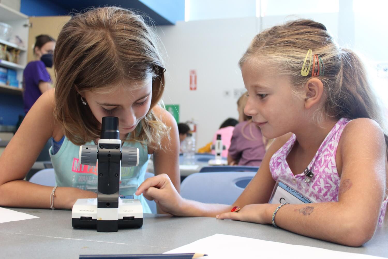 one girl looks through a microscope as another girl looks on during camps